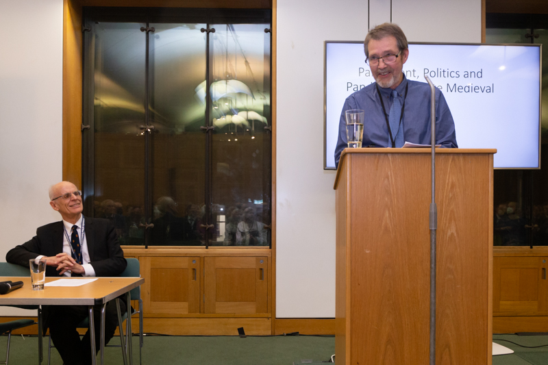 Professor Chris Given-Wilson is on the right of the image, standing behind a wooden lectern and microphone, and in front of a screen displaying the lecture title. He is wearing a grey shirt and grey tie. Lord Norton of Louth, in a white shirt, blue suit and dark tie, is on the left of the image. He is behind a table, looking towards Professor Given-Wilson.