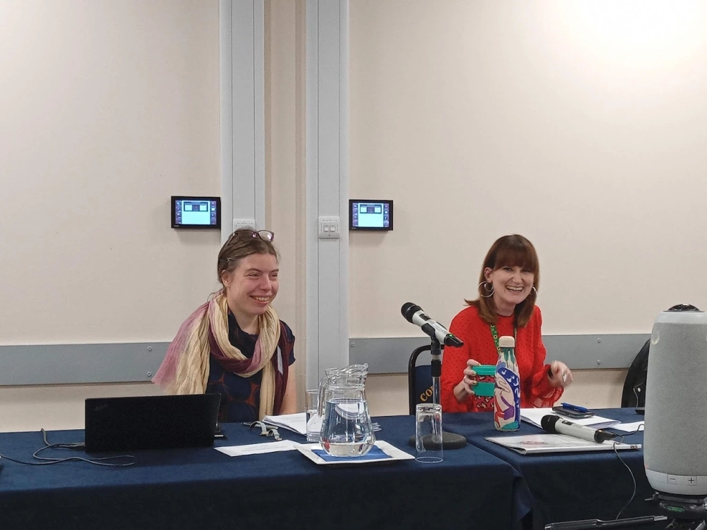 Two white women sat next to each other laughing. There is a microphone between the two of them. The woman on the left has glasses on her head, her hair back and is wearing a scarf, the woman on the right has a fringe and a red dress.